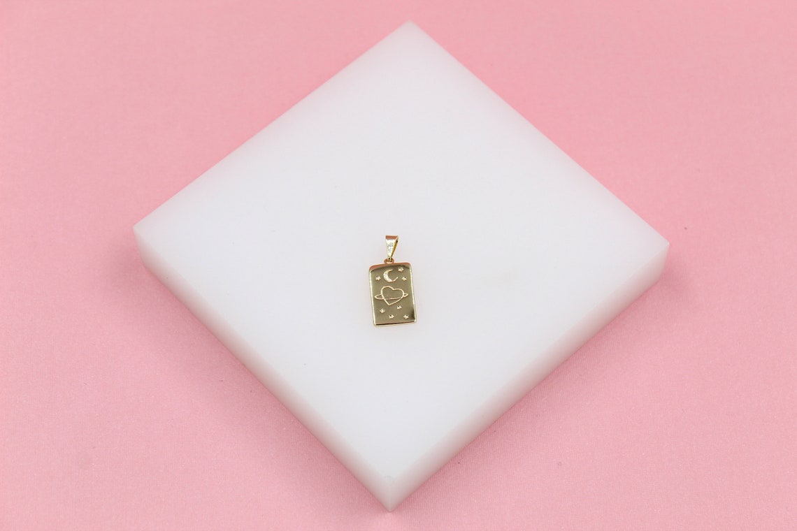 A rectangular gold pendant featuring a celestial engraved design of a crescent moon, a heart surrounded by a ring, and tiny stars rests on a white display block against a pink background.