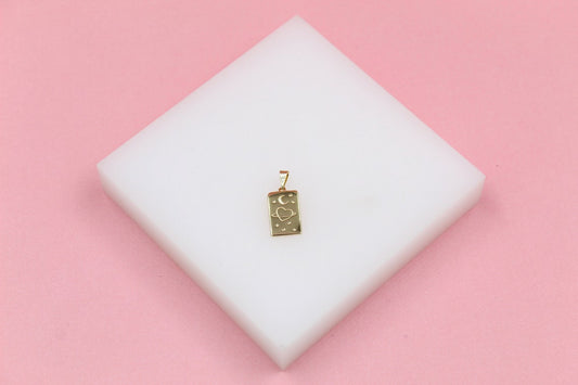 A rectangular gold pendant featuring a celestial engraved design of a crescent moon, a heart surrounded by a ring, and tiny stars rests on a white display block against a pink background.