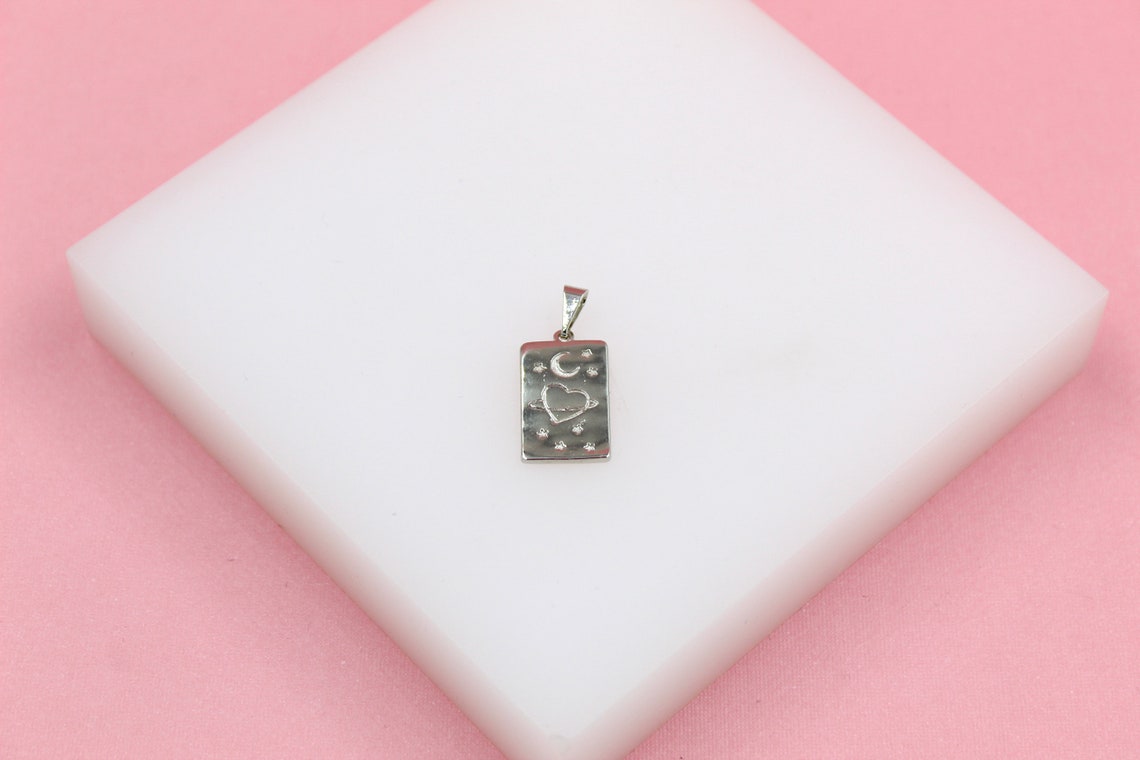 A silver celestial pendant featuring a heart, moon, and stars rests on a white display block against a pink background.