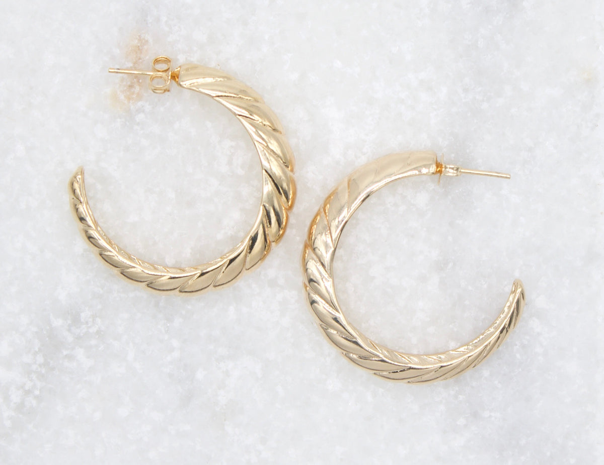 A pair of textured gold hoop earrings rests against a white marble surface.