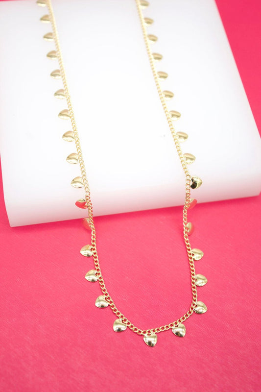A delicate gold chain necklace featuring dangling heart charms lies draped across a white display block against a vibrant pink background.