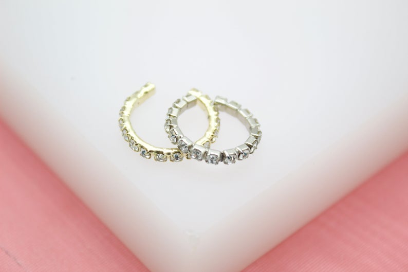 Two gold and silver rhinestone-encrusted ear cuffs rest on a white display surface against a soft pink background.