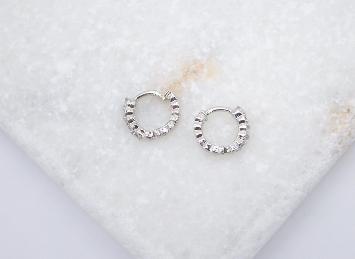 A pair of sparkling silver huggie hoop earrings rests delicately on a textured white marble surface.
