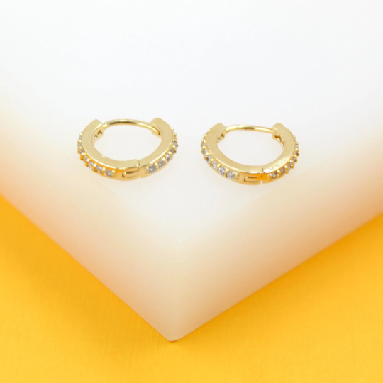 A pair of gold huggie hoop earrings embedded with sparkling cubic zirconia crystals rests on a white display block.