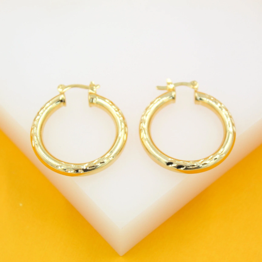 A pair of polished gold hoop earrings with a textured diamond-cut pattern rests on a white and yellow surface.