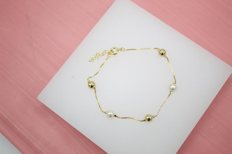 A delicate gold chain bracelet features alternating gold and pearl beads, resting on a white display surface.