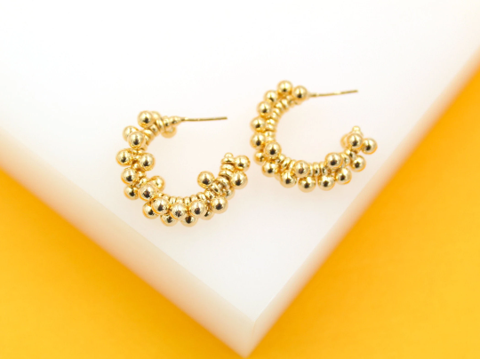 Two gold beaded hoop earrings rest on a white display surface against a vibrant yellow background.