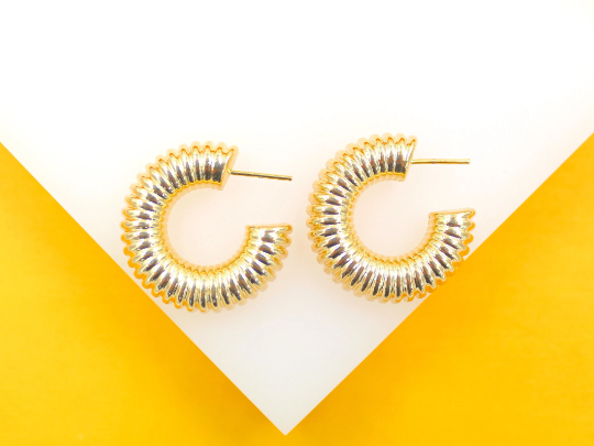 A pair of thick gold hoop earrings with a ribbed texture rest on a white surface against a bright yellow background.