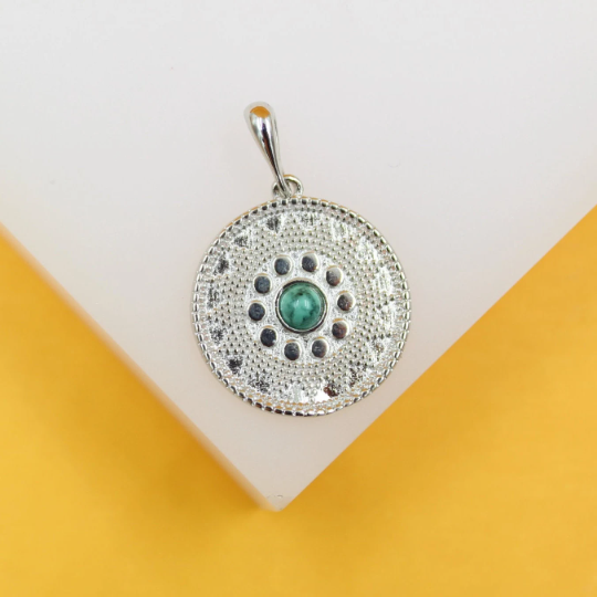 A silver pendant necklace features an intricate circular coin design centered with a vibrant green gemstone.