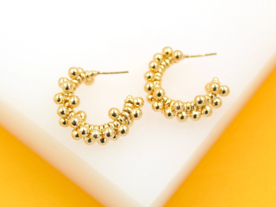 A pair of textured gold half-hoop earrings featuring clustered bead details rests on a white and yellow surface.