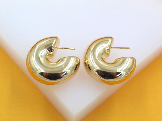 A pair of chunky gold hoop earrings rests neatly on a white display surface against a vibrant orange background.