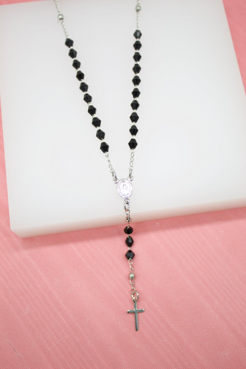 A silver rosary necklace features faceted black crystal beads and a matching crucifix pendant displayed against a pink background.