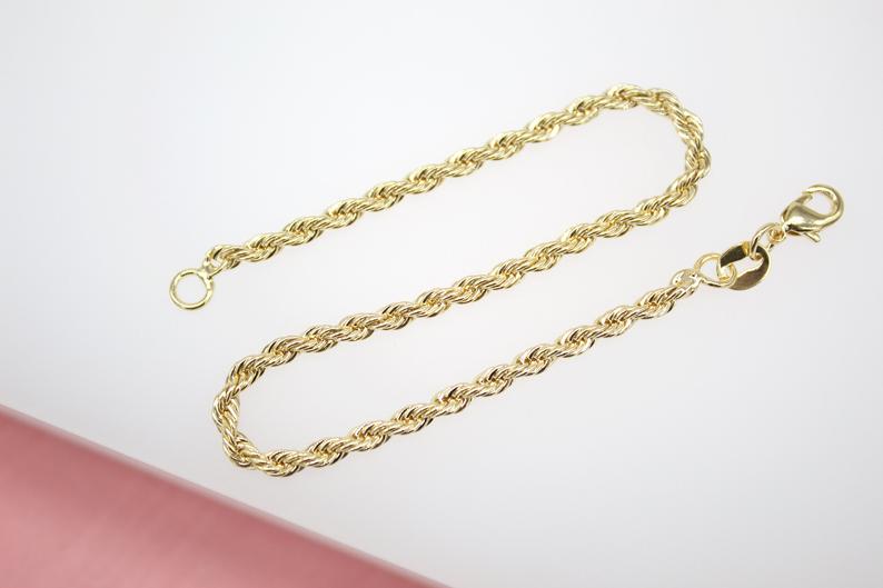 A shiny gold rope chain bracelet lies against a neutral white background next to a pink surface.