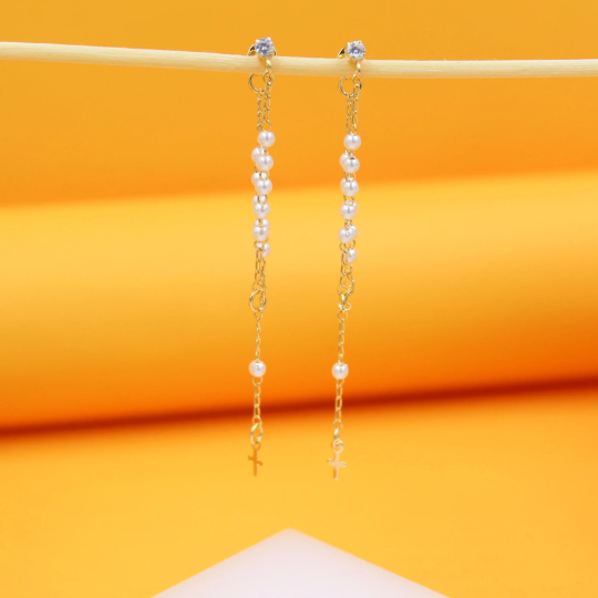 A pair of delicate gold chain earrings featuring dangling pearls and small cross charms hang against an orange backdrop.