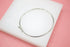 A sleek silver bangle bracelet rests on a white display block against a soft pink background.
