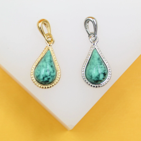 Two teardrop-shaped turquoise gemstone pendants, one in a gold setting and the other in silver, rest side by side on a white display against a yellow background.