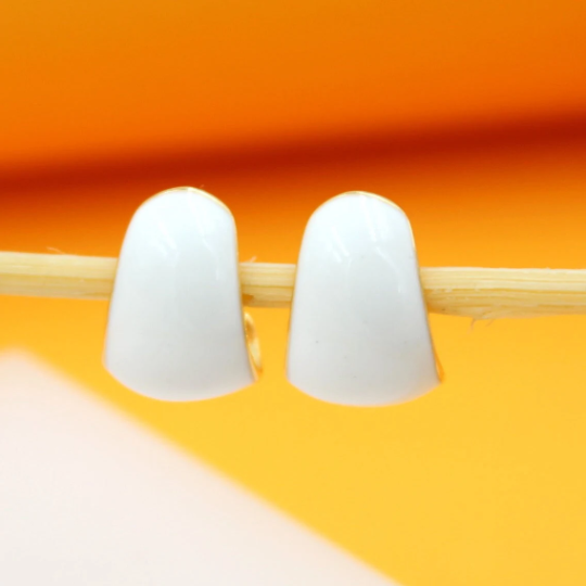 A pair of glossy white huggie hoop earrings rests against a wooden stick against an orange background.