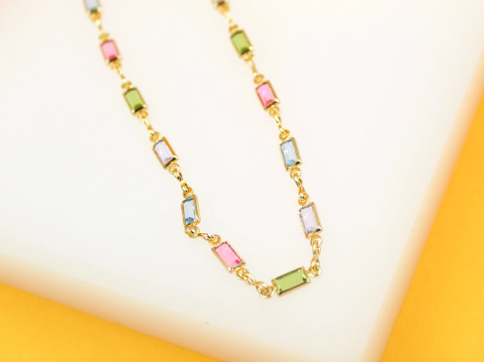 A delicate gold necklace features colorful baguette-cut stones in pink, green, and blue displayed against a white and yellow background.
