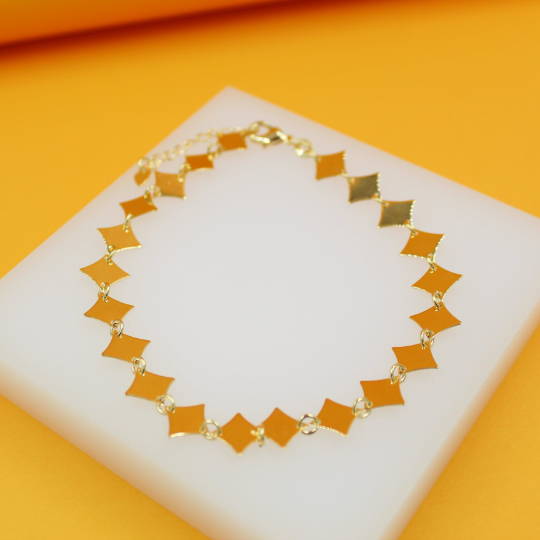 A delicate gold bracelet featuring geometric diamond-shaped charms rests on a white display block.