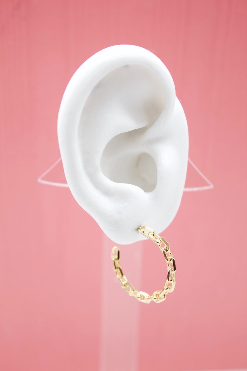 A white mannequin ear displays a gleaming gold chain-link hoop earring against a pink background.