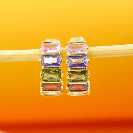 A pair of gold huggie hoop earrings featuring a colorful array of baguette-cut pink, purple, green, and red cubic zirconia stones rests on a wooden stick against a warm yellow background.
