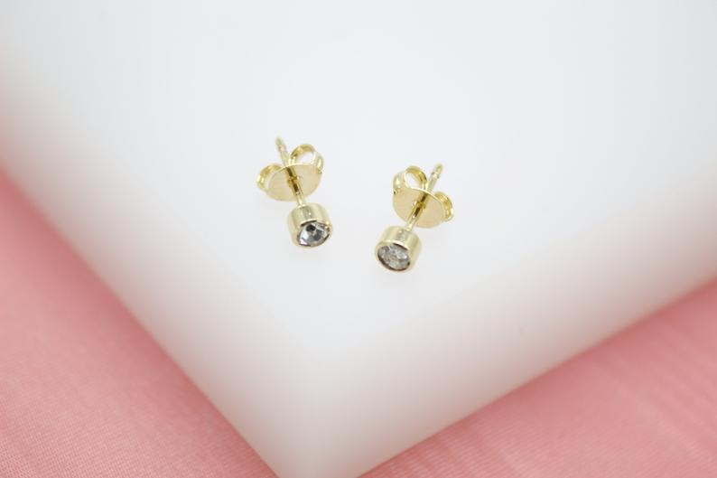 A pair of gold bezel-set crystal stud earrings rest on a white surface against a pink background.