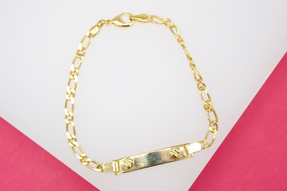 A polished gold-plated identification bracelet rests on a white and pink background.