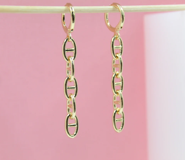 Two gleaming gold chain-link dangle earrings hang side-by-side against a soft pink backdrop.
