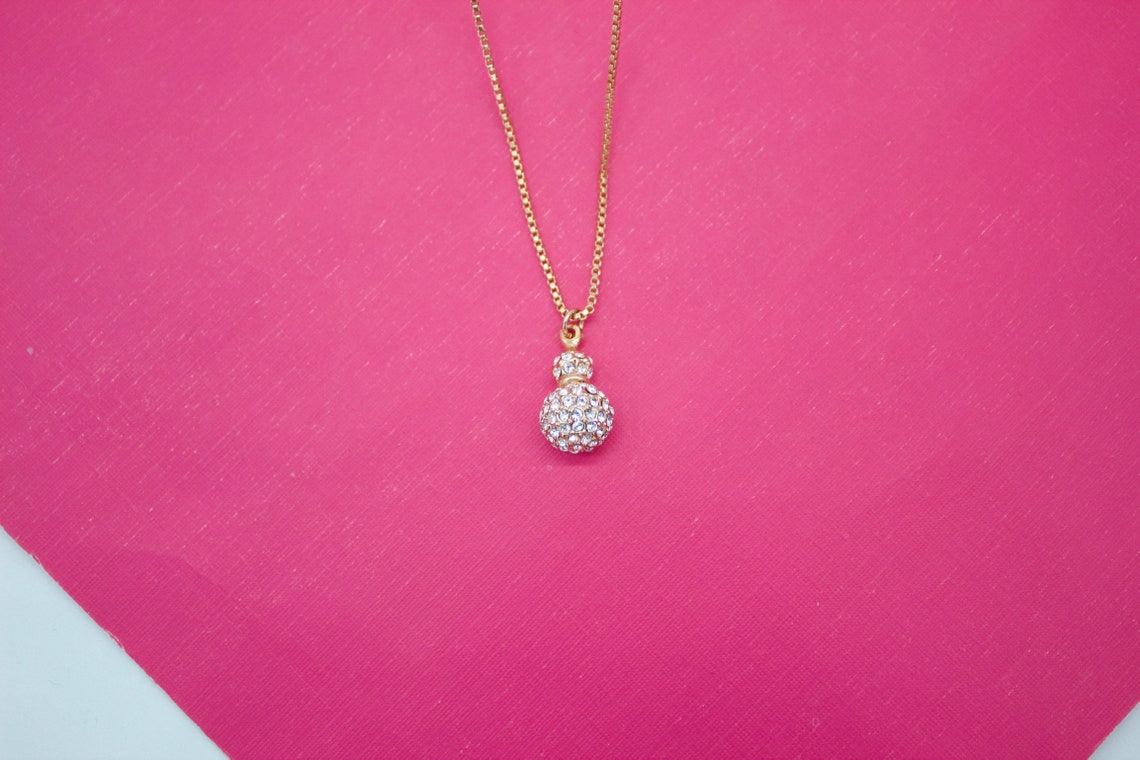 A delicate gold necklace with a rhinestone-encrusted sphere pendant rests against a vibrant pink background.