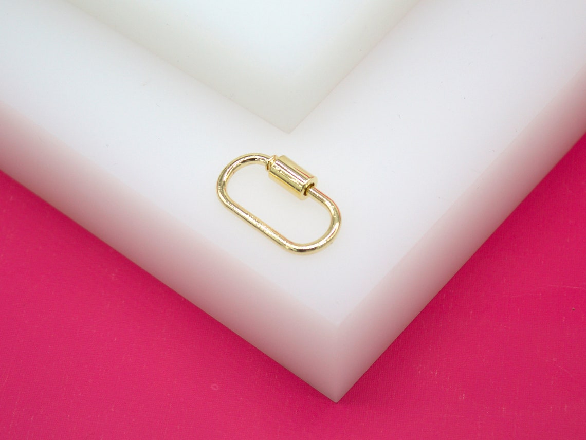 A shiny gold screw-lock carabiner charm rests on a white block against a bright pink background.