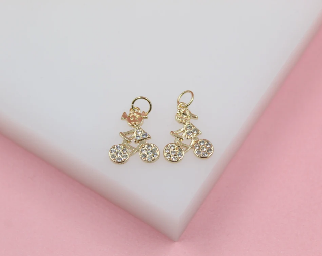 Two gold bicycle charms embellished with sparkling cubic zirconia stones rest on a white display surface against a pink background.