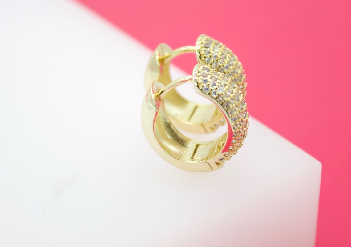 A pair of gold huggie hoop earrings encrusted with sparkling cubic zirconia stones rests on a white display surface against a pink background.
