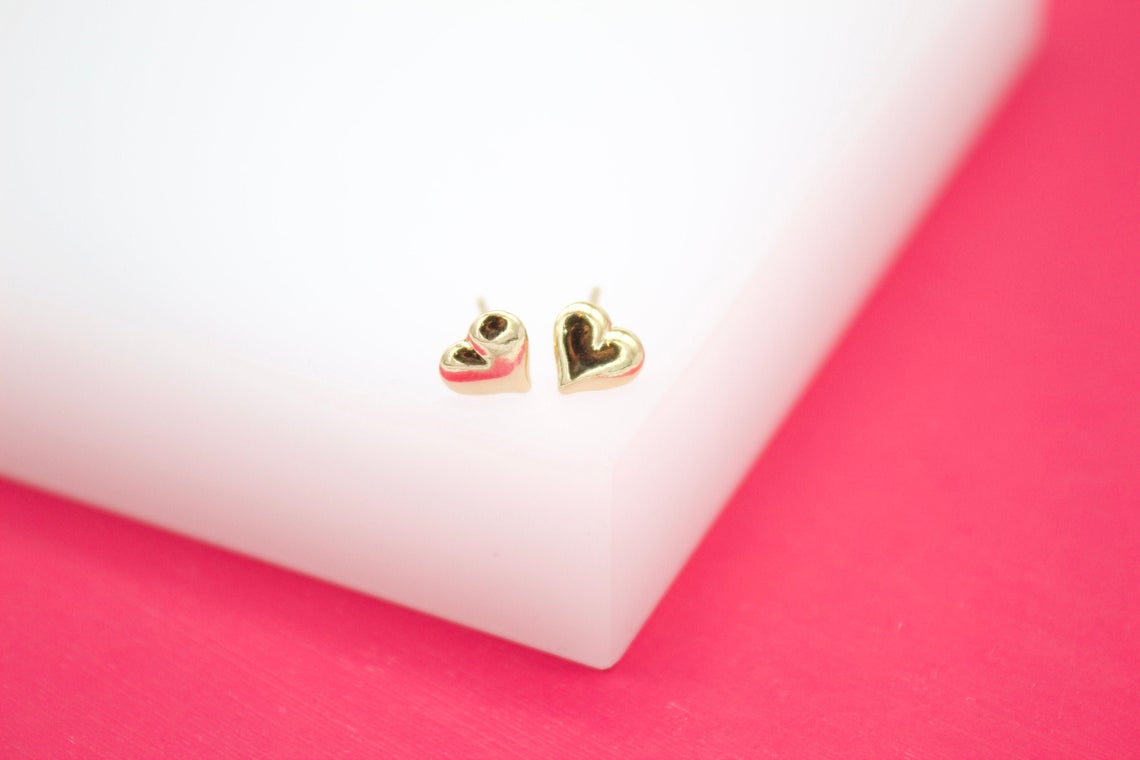 A pair of dainty gold heart stud earrings rests on a white block against a vibrant pink background.