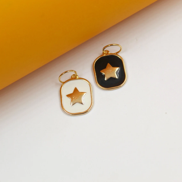 Two rectangular gold-rimmed enamel pendants showcase a white and a black star charm resting side by side against a yellow and white background.