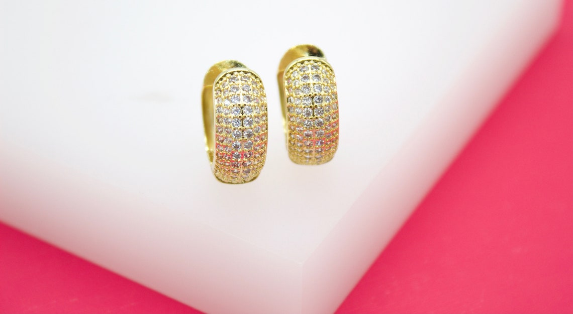 A pair of gold huggie hoop earrings encrusted with sparkling cubic zirconia stones rests on a white display block against a vibrant pink background.