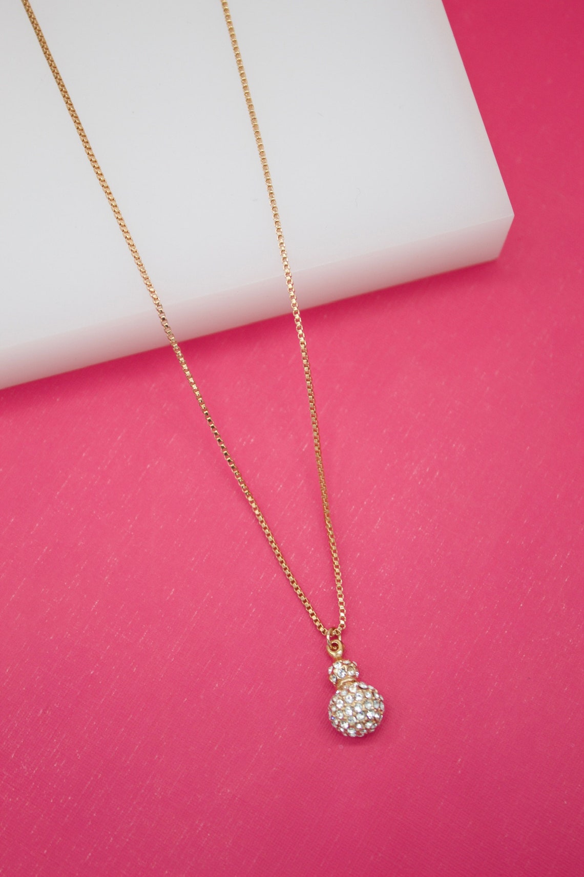 A delicate gold chain necklace hangs against a vibrant pink backdrop, featuring a sparkling pave crystal gourd pendant.
