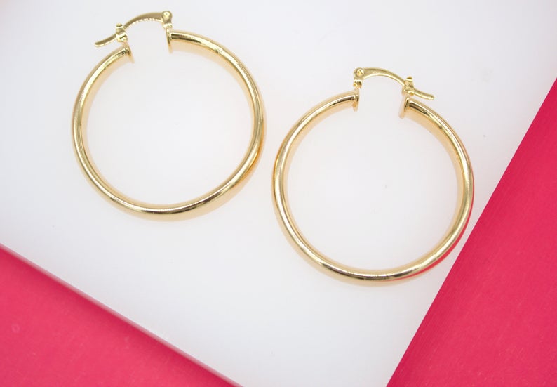 Two polished gold hoop earrings rest on a white surface against a vibrant pink background.