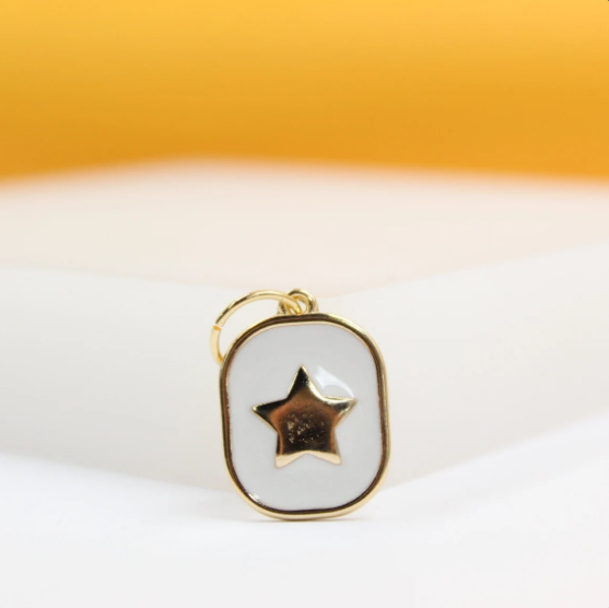 A gold-rimmed rectangular pendant with white enamel features a shiny gold star in the center against a soft-focus background.