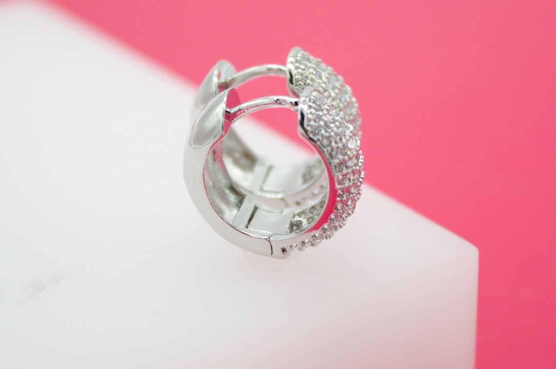 A sparkling silver huggie hoop earring encrusted with cubic zirconia stones rests upright on a white and pink surface.