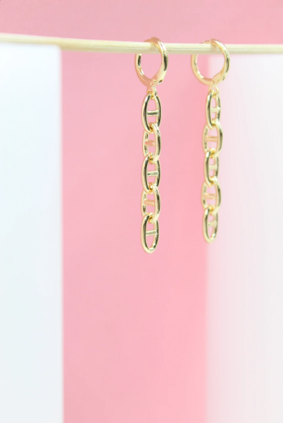 A pair of elegant gold chain link dangle earrings hangs from a stick against a pink and white backdrop.