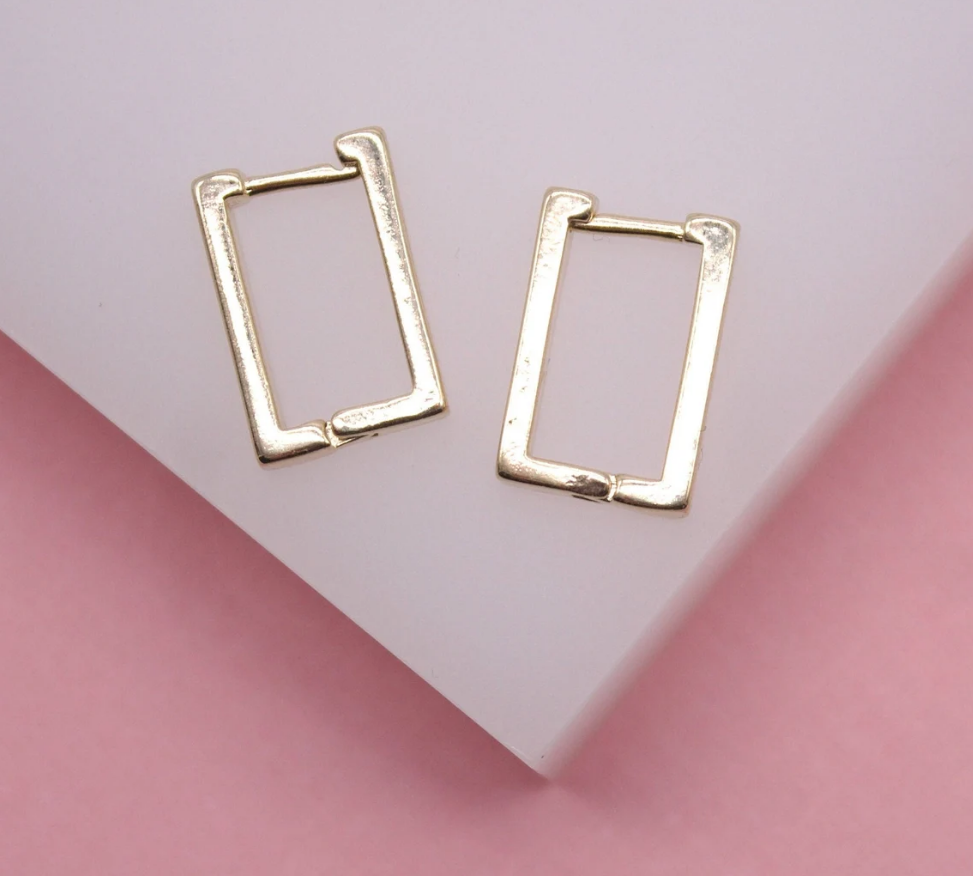A pair of gold rectangular hoop earrings rests on a white display surface against a pink background.