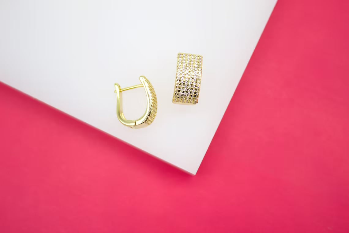 A pair of sparkling pave-set gold hoop earrings rests on a white display against a vibrant pink background.