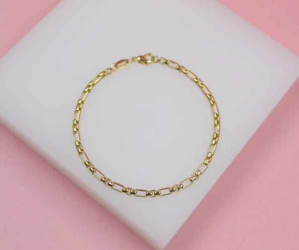 A delicate gold chain bracelet rests centered on a white display pad against a soft pink background.