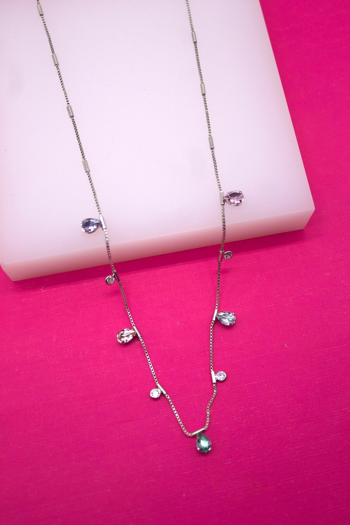 A delicate silver chain necklace featuring colorful dangling teardrop and round cubic zirconia stones rests on a white display block against a vibrant pink background.