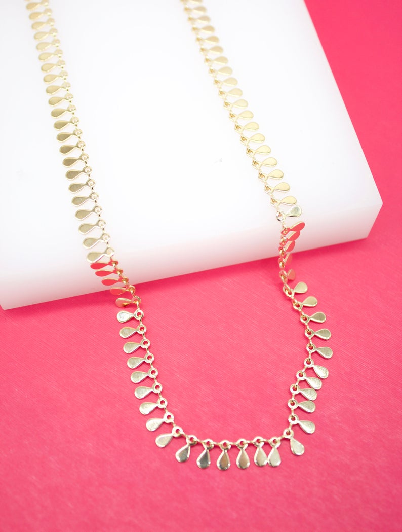 A delicate gold teardrop station necklace rests on a white display block against a vibrant pink background.