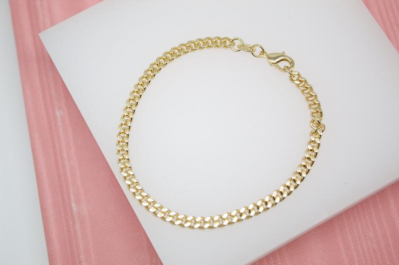 A gleaming gold cuban link chain bracelet rests gracefully on a white display surface against a pink background.
