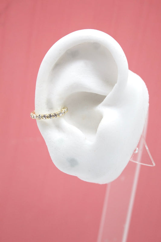 A white mannequin ear displays a sparkling gold ear cuff encrusted with tiny gemstones against a soft pink background.