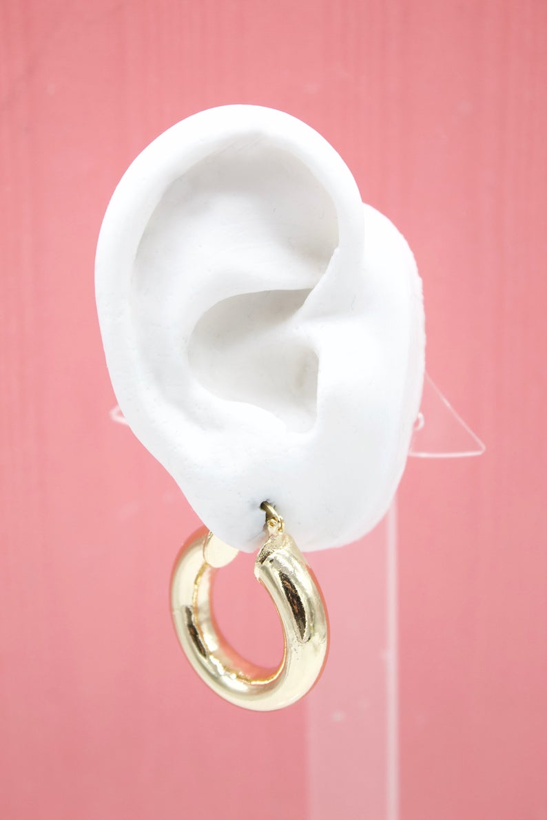 A thick gold hoop earring hangs from a white display ear against a solid pink background.