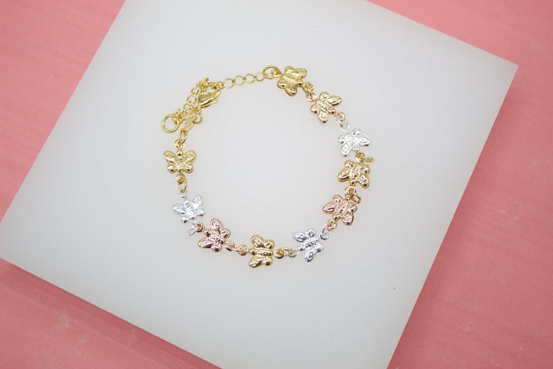 A delicate gold tri-color butterfly bracelet rests on a white display surface against a pink background.