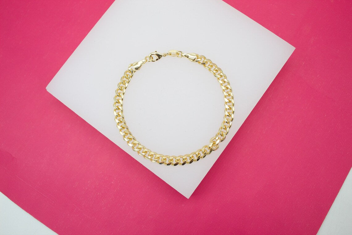 A polished gold cuban link bracelet rests in a circle on a white platform against a vibrant pink background.